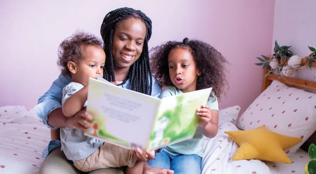 Marielle Altenor sits on a white polka dot bed reading a story book with her little son and daughter on her lap.