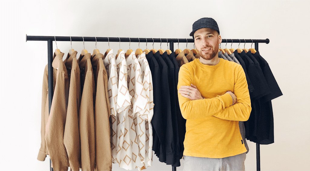 A smiling Matteo wearing a yellow sweater and ballcap stands in front of a clothing rack.