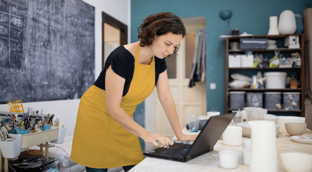 Une femme qui porte un tablier jaune utilise un ordinateur portable dans l’atelier d’une petite entreprise.