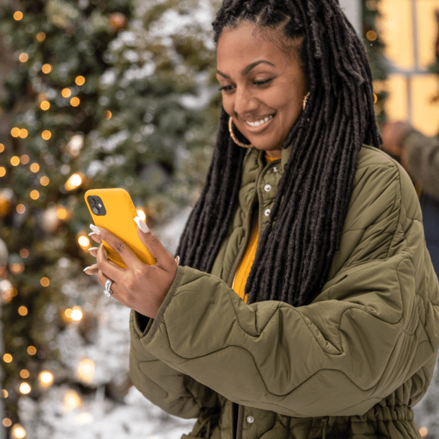 Woman in green coat looks down at her yellow phone and smiles as people decorate behind her.