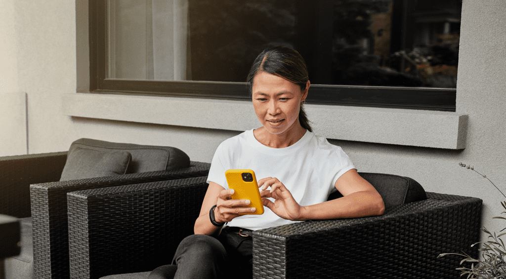 Woman sits on her deck using her cellphone to make an Interac e-Transfer for Business transaction.