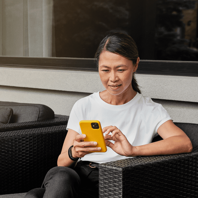 Woman sits on her deck using her cellphone to make an Interac e-Transfer for Business transaction.